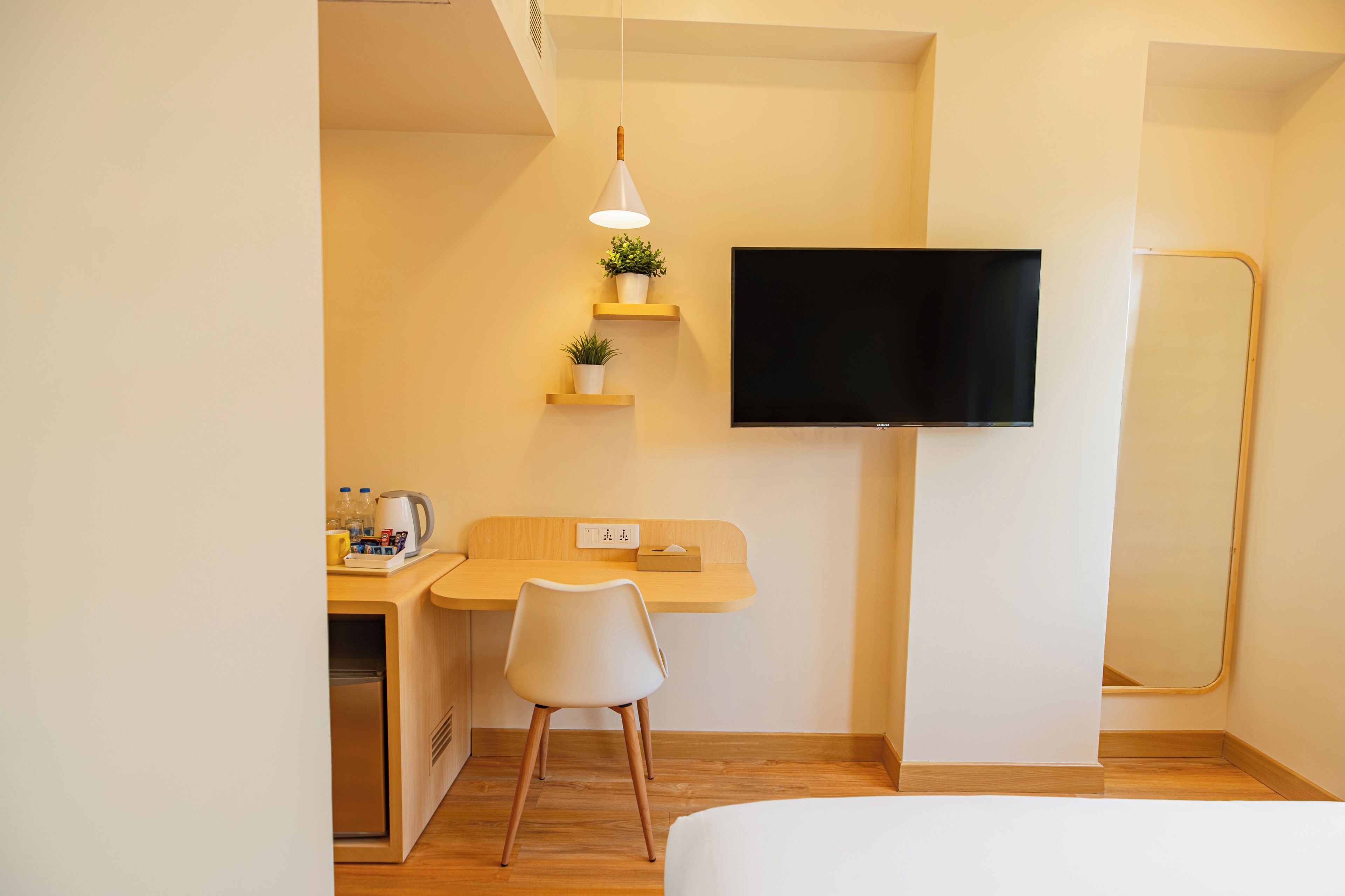 A hotel room with a bed, a desk with a chair, a TV mounted on the wall, and a small shelf with plants.
