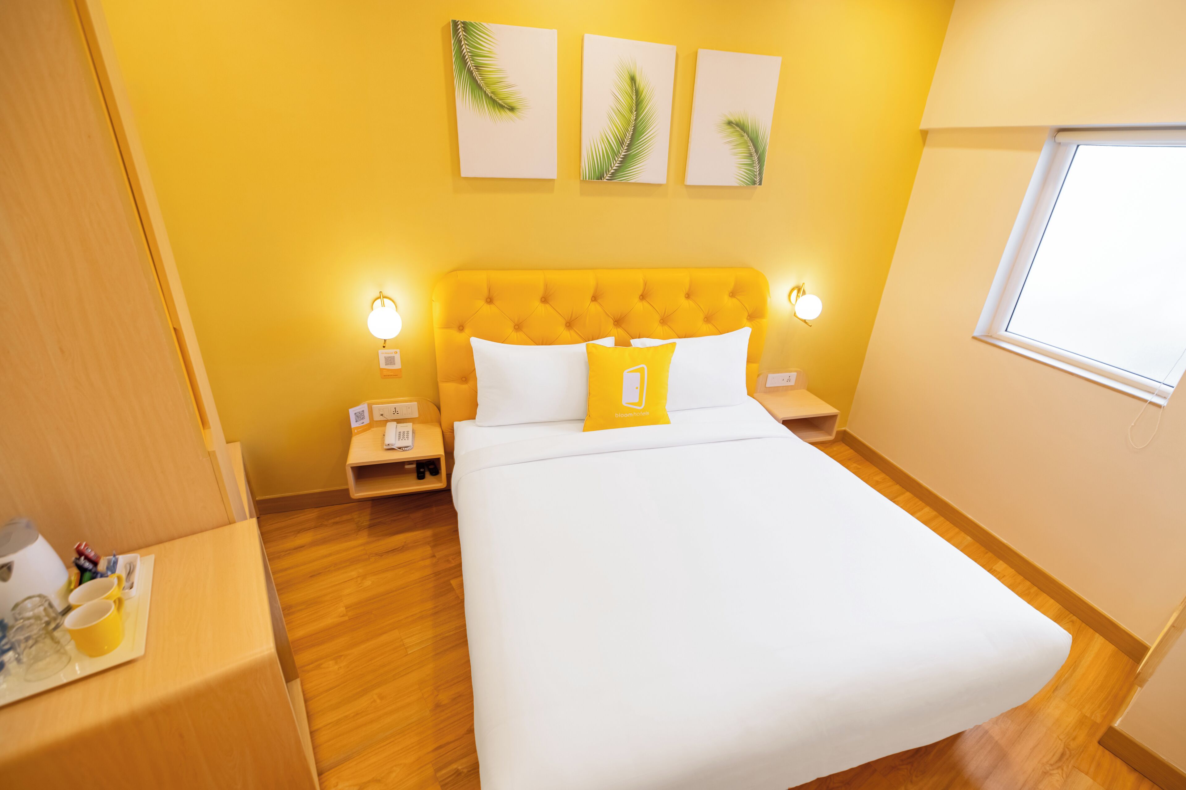 Una habitación de hotel con pared amarilla, cabecera capitonada, ropa de cama blanca y una ventana.