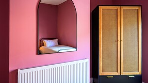 Une pièce avec un mur rose, un radiateur blanc, un lit avec un oreiller blanc et une armoire en bois à deux portes.