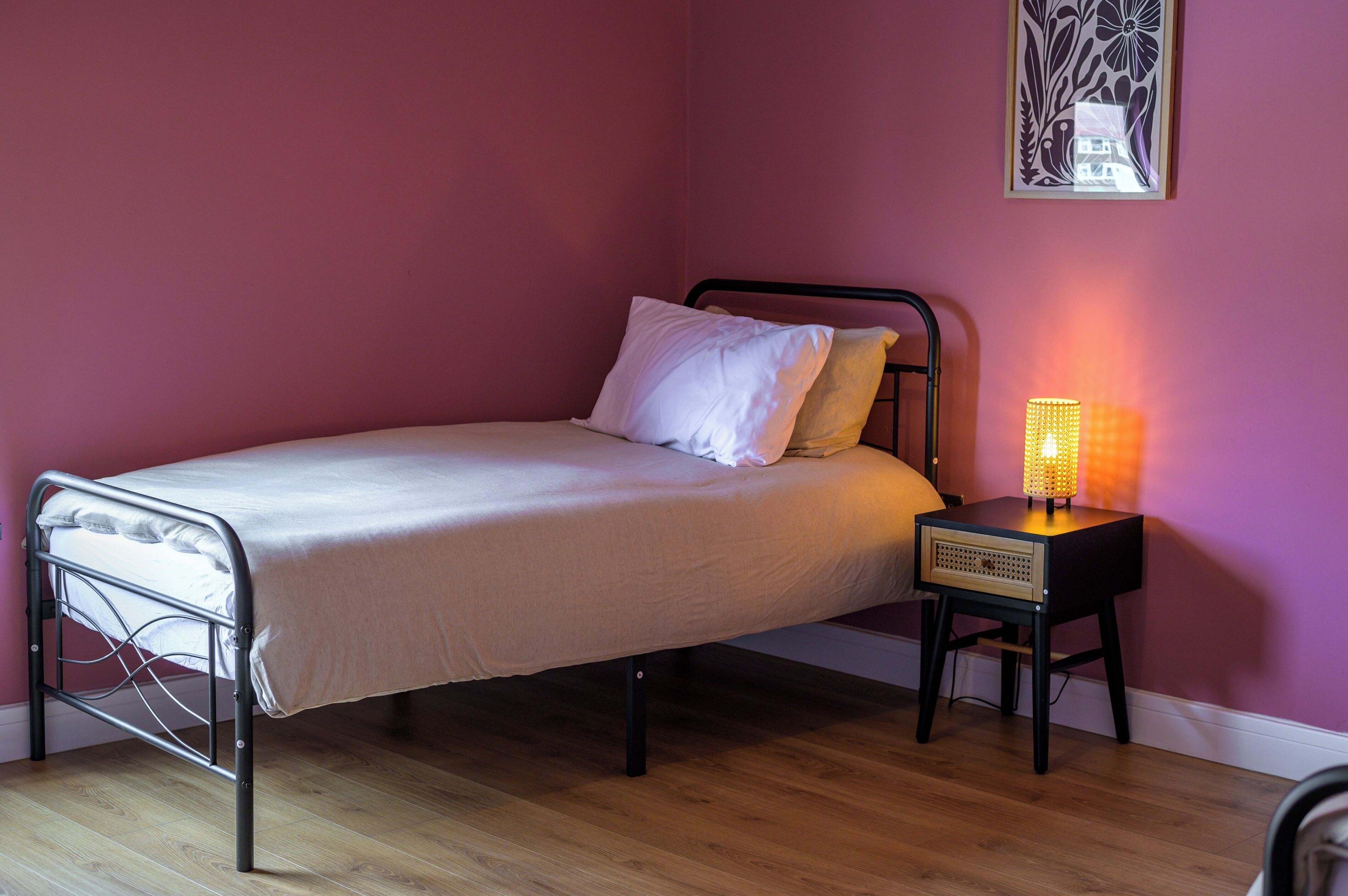 A single bed with a metal frame, a bedside table with a lamp, and a wall-mounted artwork.