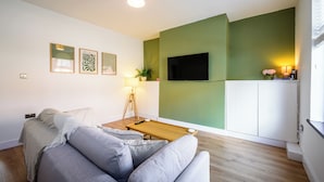Un salon moderne avec un canapé gris, une table basse en bois, un téléviseur à écran plat fixé sur un mur vert et un meuble blanc.