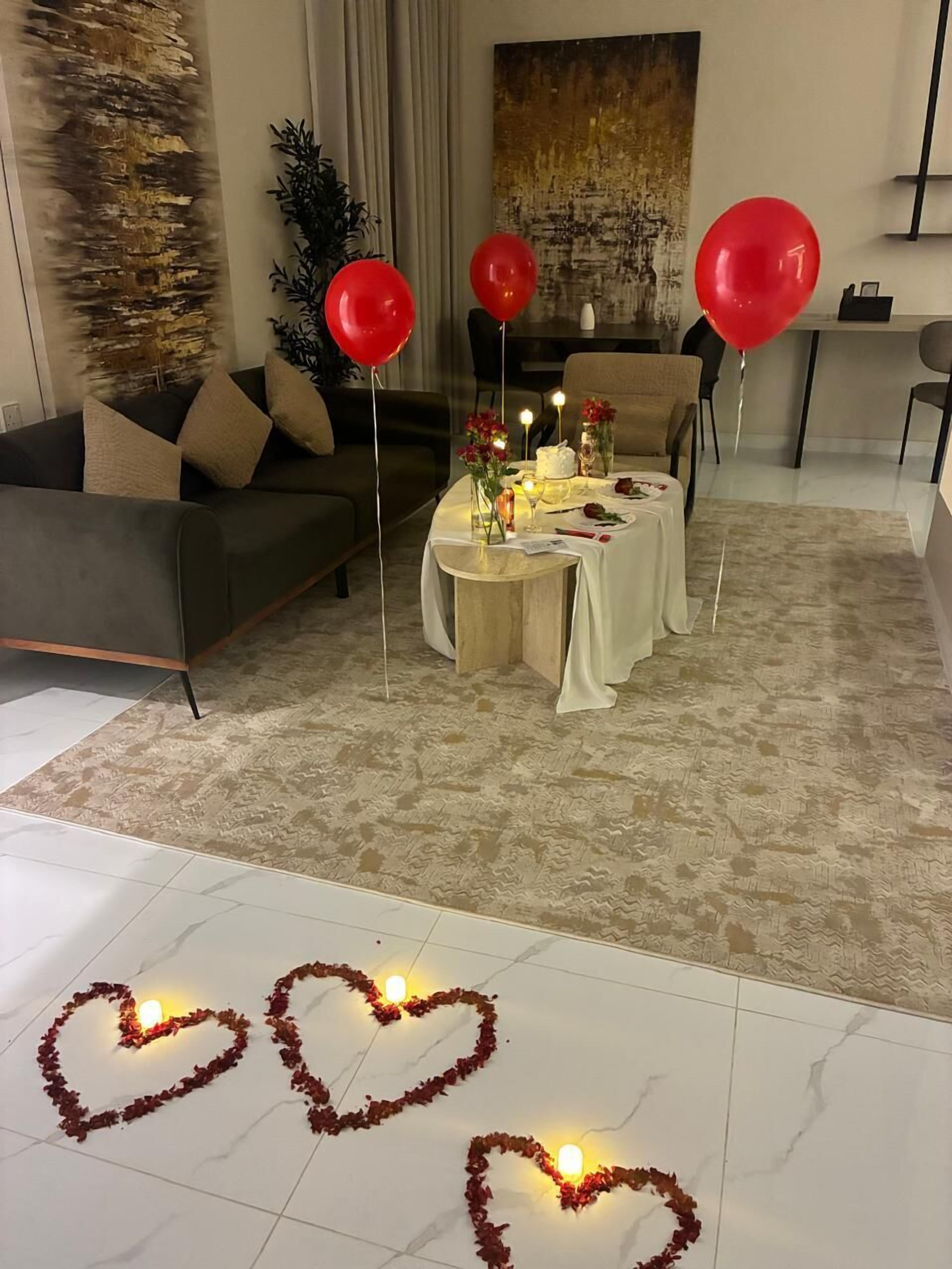 Una sala de estar con un sofá, una mesa de comedor con pastel y velas, y arreglos florales con forma de corazón en el suelo.