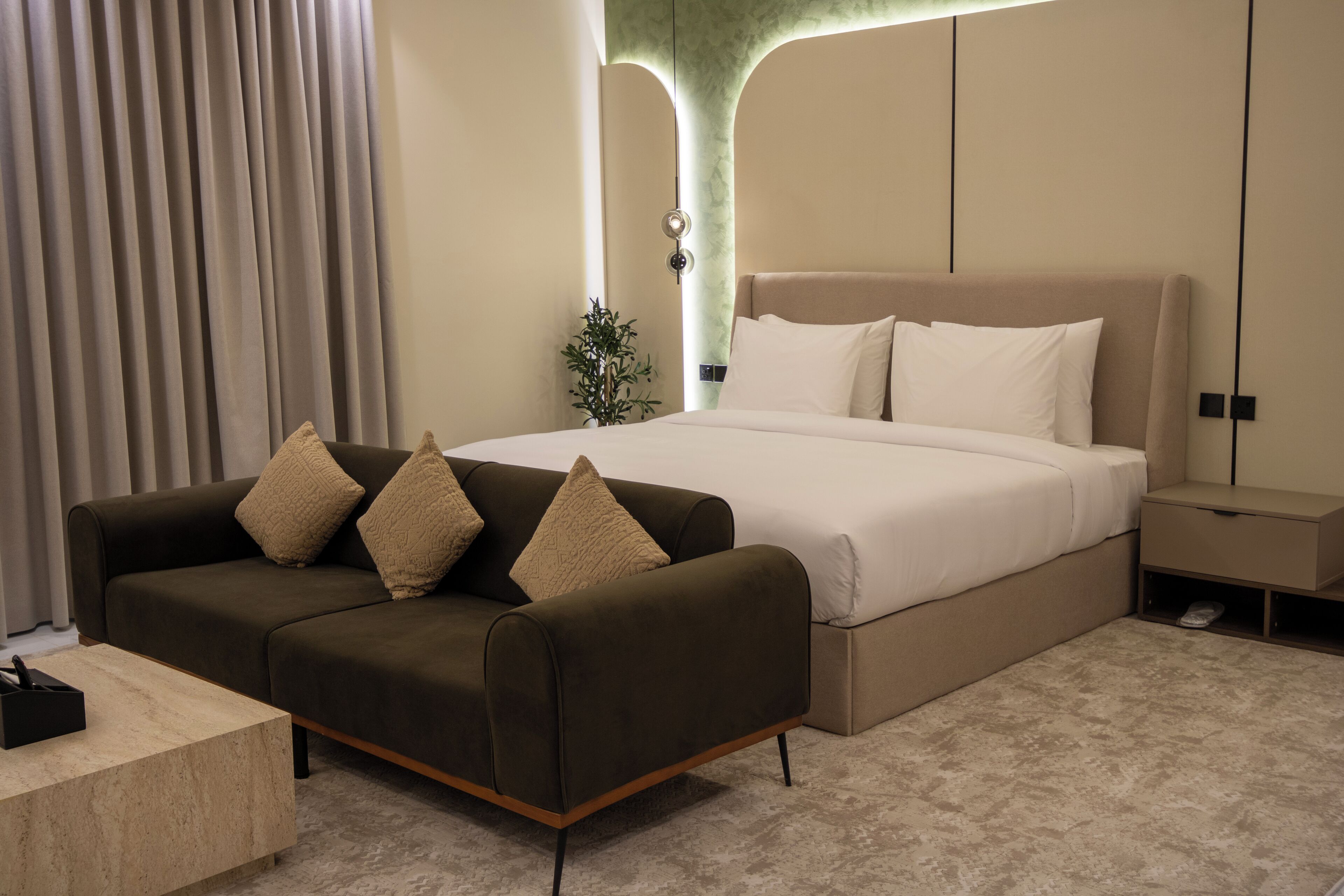 A modern bedroom with a large bed, a dark sofa with cushions, a bedside table, and a small plant.
