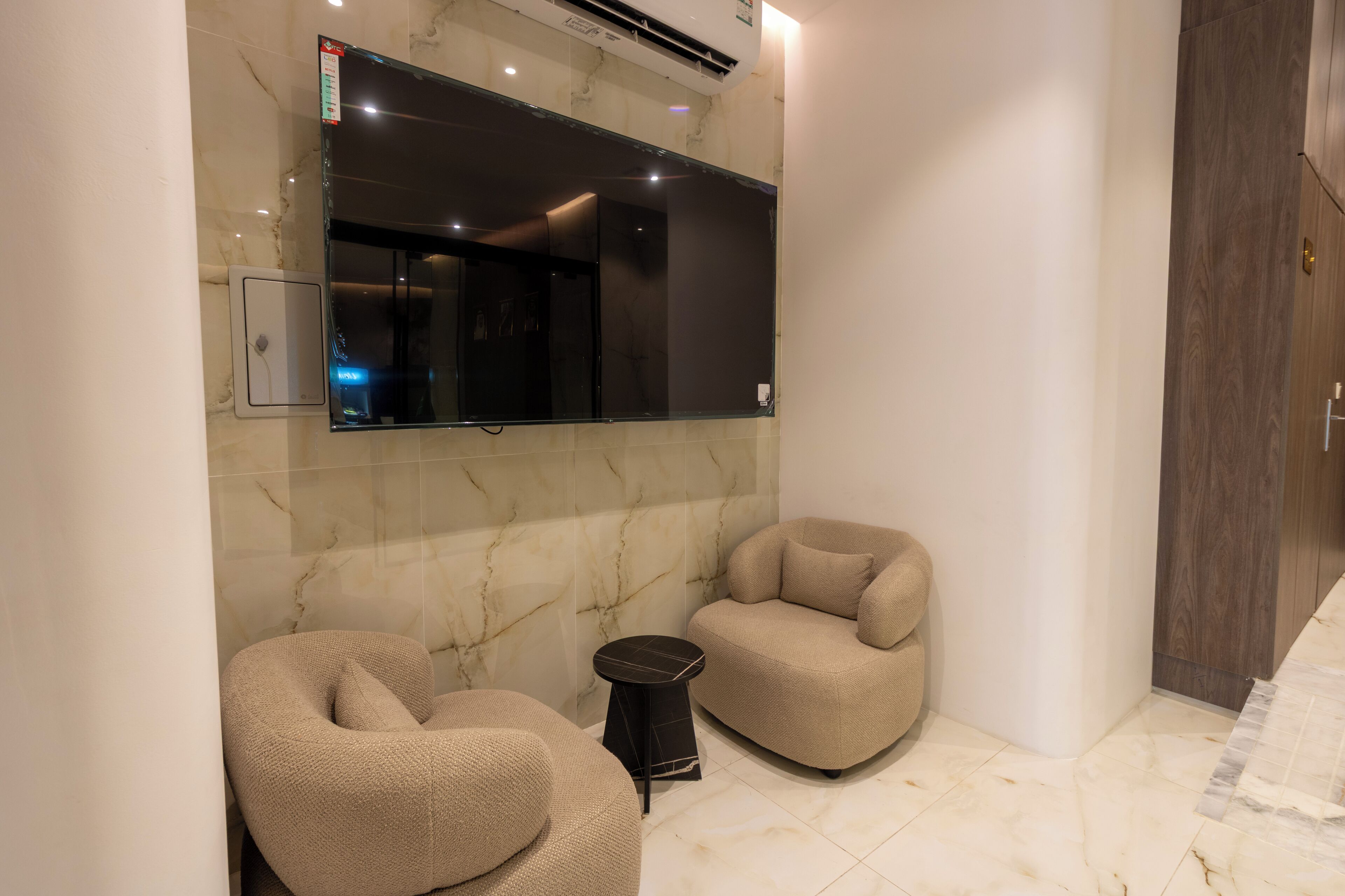 A modern bathroom with a large mirror, two beige chairs, a small round table, and a marble floor.
