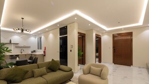 A modern living room with a sofa, armchair, and coffee table. There is a kitchen area with a dining table and chairs. The room has a wooden door and a ceiling light fixture. - Luxury suites Hotel Apartments (Taif)