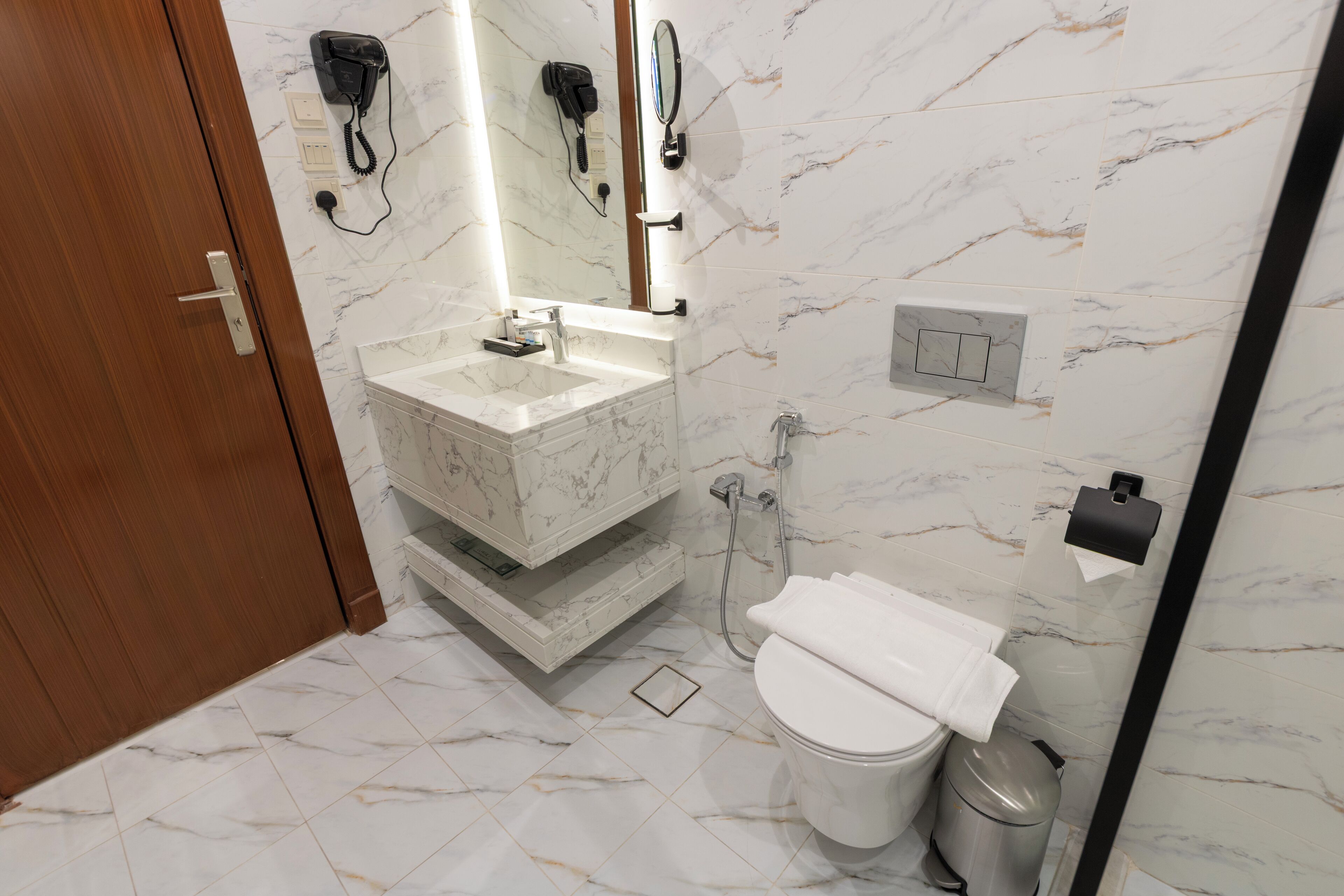 A bathroom with a marble sink, a toilet, a trash can, and a hairdryer mounted on the wall.