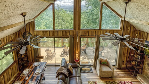 A spacious loft with a high ceiling, wooden walls, and large windows offering a view of the outdoors. There is a dining area with a long table set for four, a leather sofa, and a small armchair. Two ceiling fans are visible. - Pigeon Forge Mountain View Cabin Rental (Pigeon Forge)