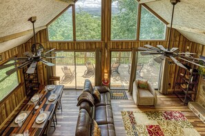 Un loft spacieux avec de hauts plafonds, des murs en bois et de grandes fenêtres offrant une vue sur l’extérieur. On y trouve un coin repas avec une longue table dressée pour quatre personnes, un canapé en cuir et un petit fauteuil. Deux ventilateurs de plafond sont visibles.
