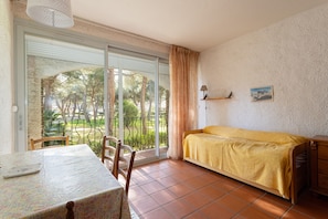 A room with a bed, a table, chairs, a lamp, a picture on the wall, and a view of trees through a sliding door. - Argelès Plage Apartment – Mountain View & Balcony, 300 m from the Beach (Argelès-sur-Mer)