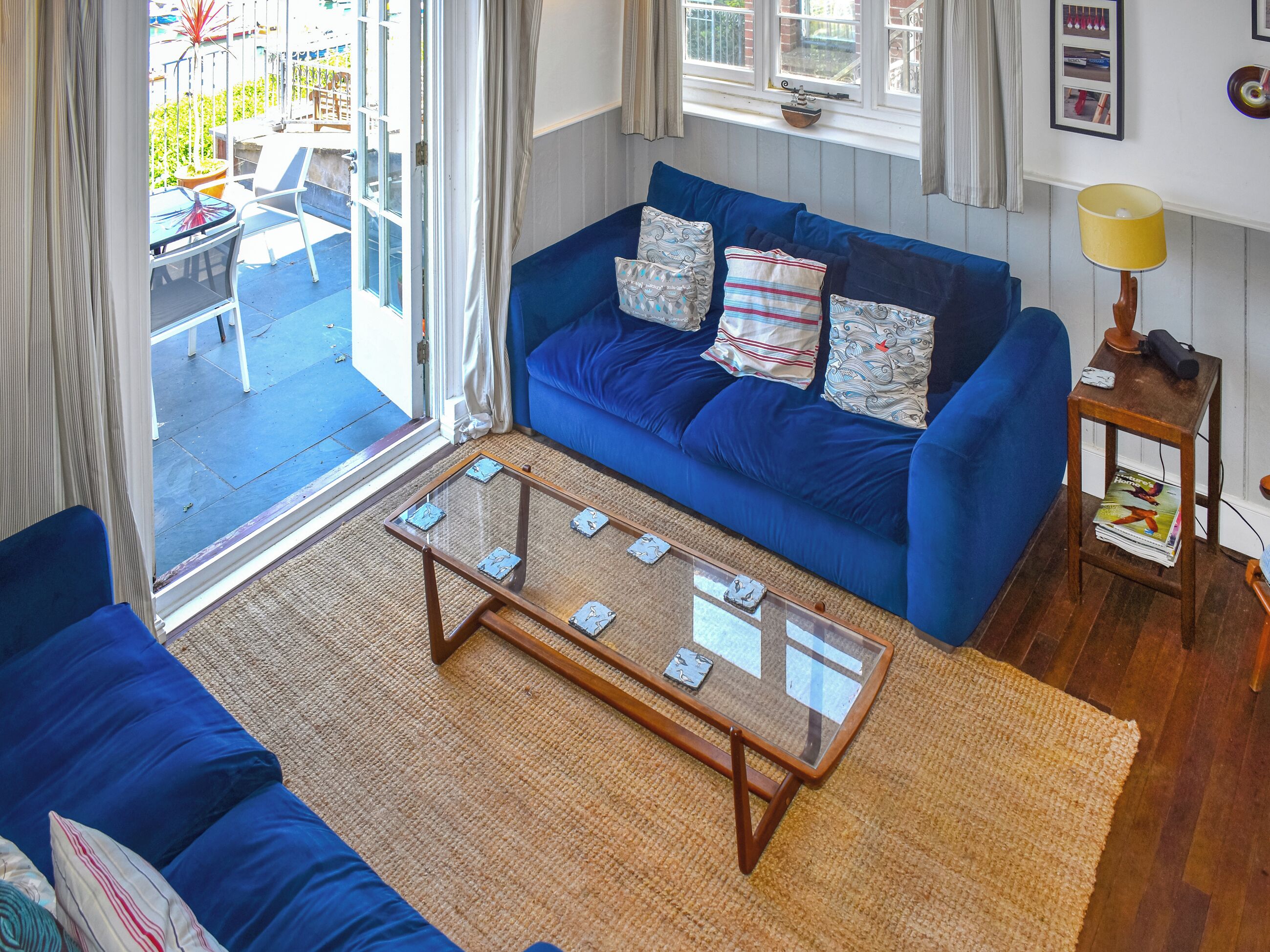 Uma sala de estar com um sofá azul, uma mesa de centro de vidro e uma pequena mesa lateral de madeira com um abajur.