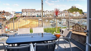 Ein Balkon mit Tisch und Stühlen, der Blick auf einen Strand und einen Yachthafen mit Booten und Gebäude im Hintergrund bietet.
