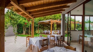 Zona pranzo all'aperto con tavoli e sedie, coperta da una pergola in legno, immersa nel verde.