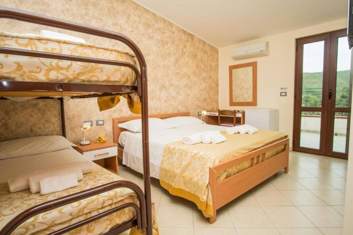 A hotel room with a bunk bed, a single bed, a desk with a chair, a mirror, and a window with a view of greenery.