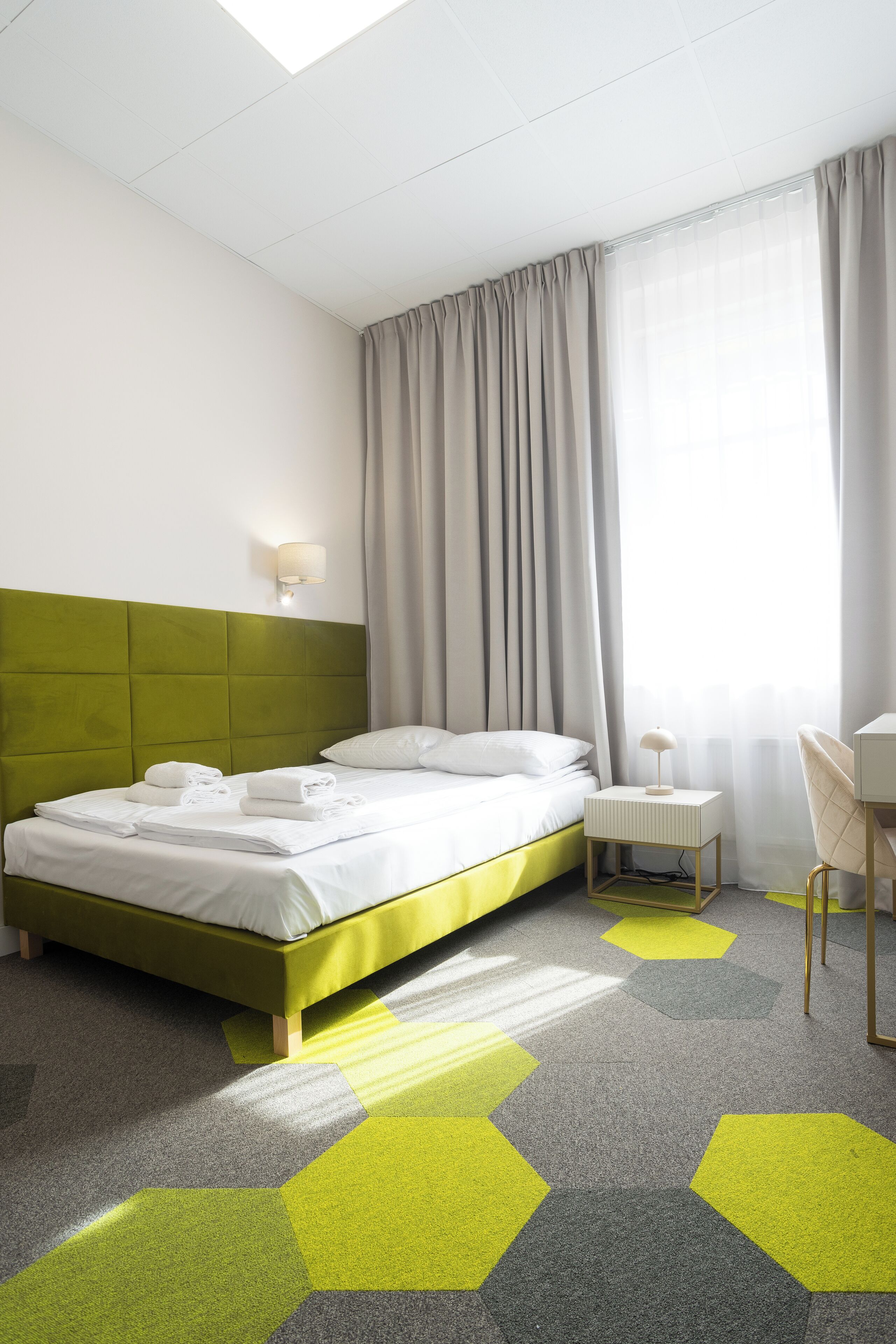 A hotel room with a green bed, white linens, a small bedside table with a lamp, a chair, and a window with curtains.
