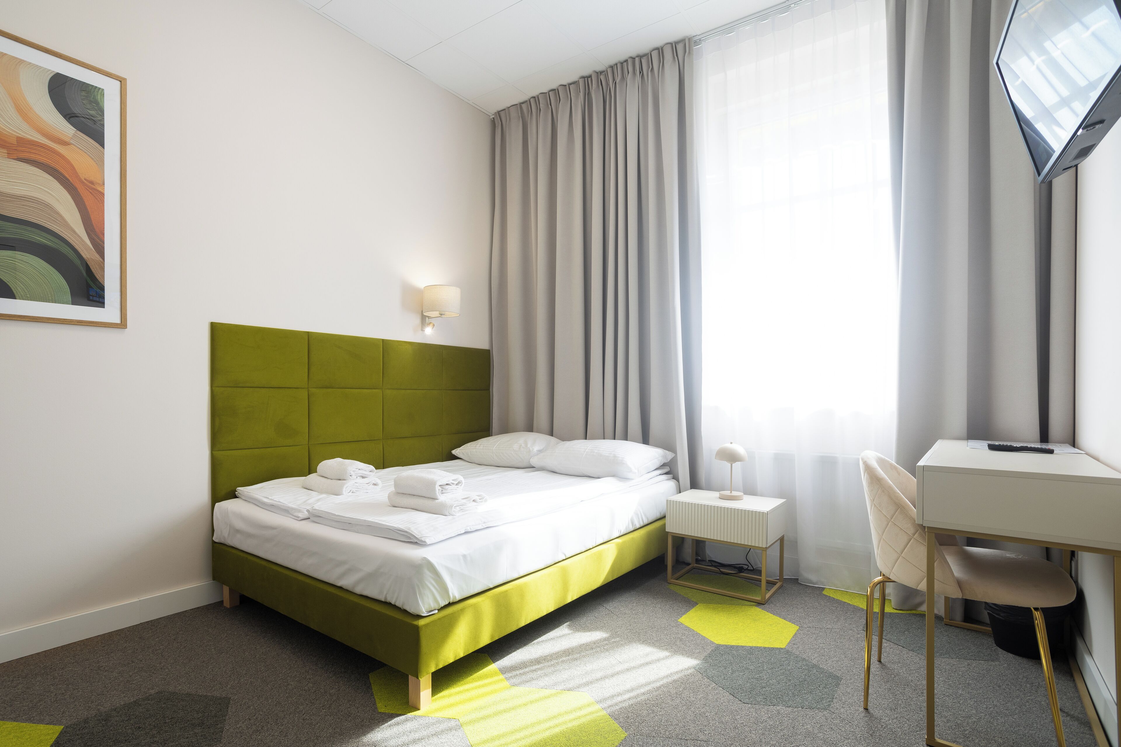 Een hotelkamer met een met groen bekleden bed, witte lakens, een bureau met stoel, een flatscreen-tv en een schilderij aan de muur.