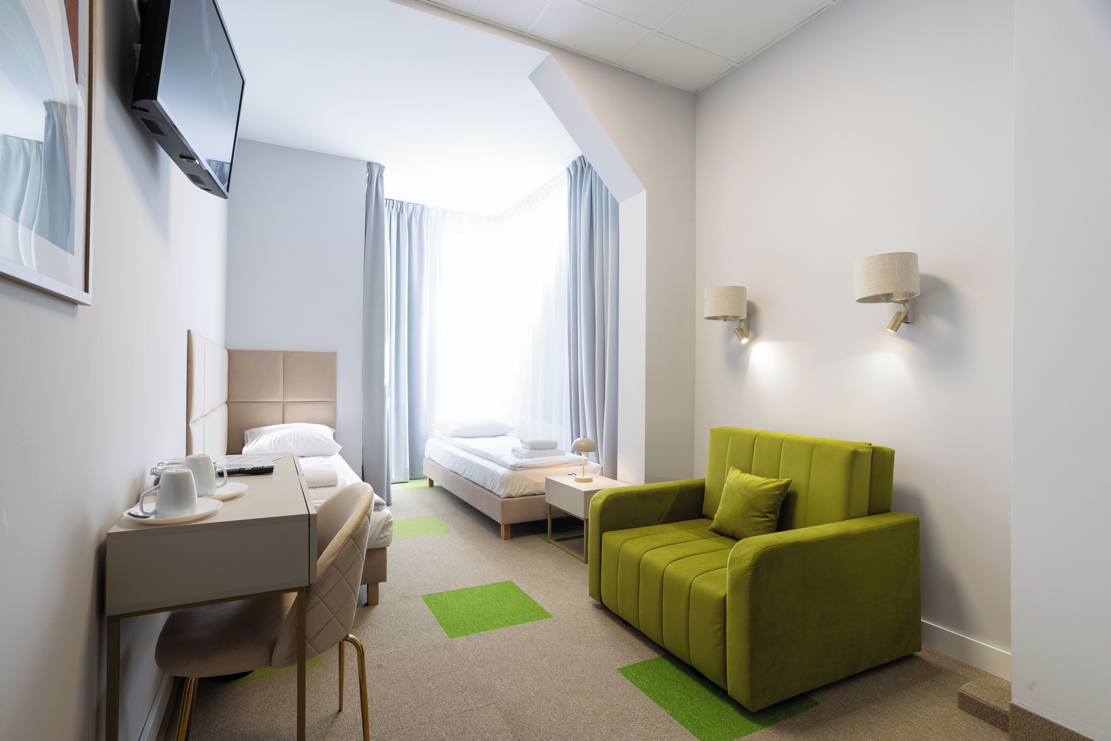 Hotellihuone, jossa on sänky, työpöytä tuolilla, vihreä sohva, seinälle asennettu televisio ja kaksi seinävalaisinta.