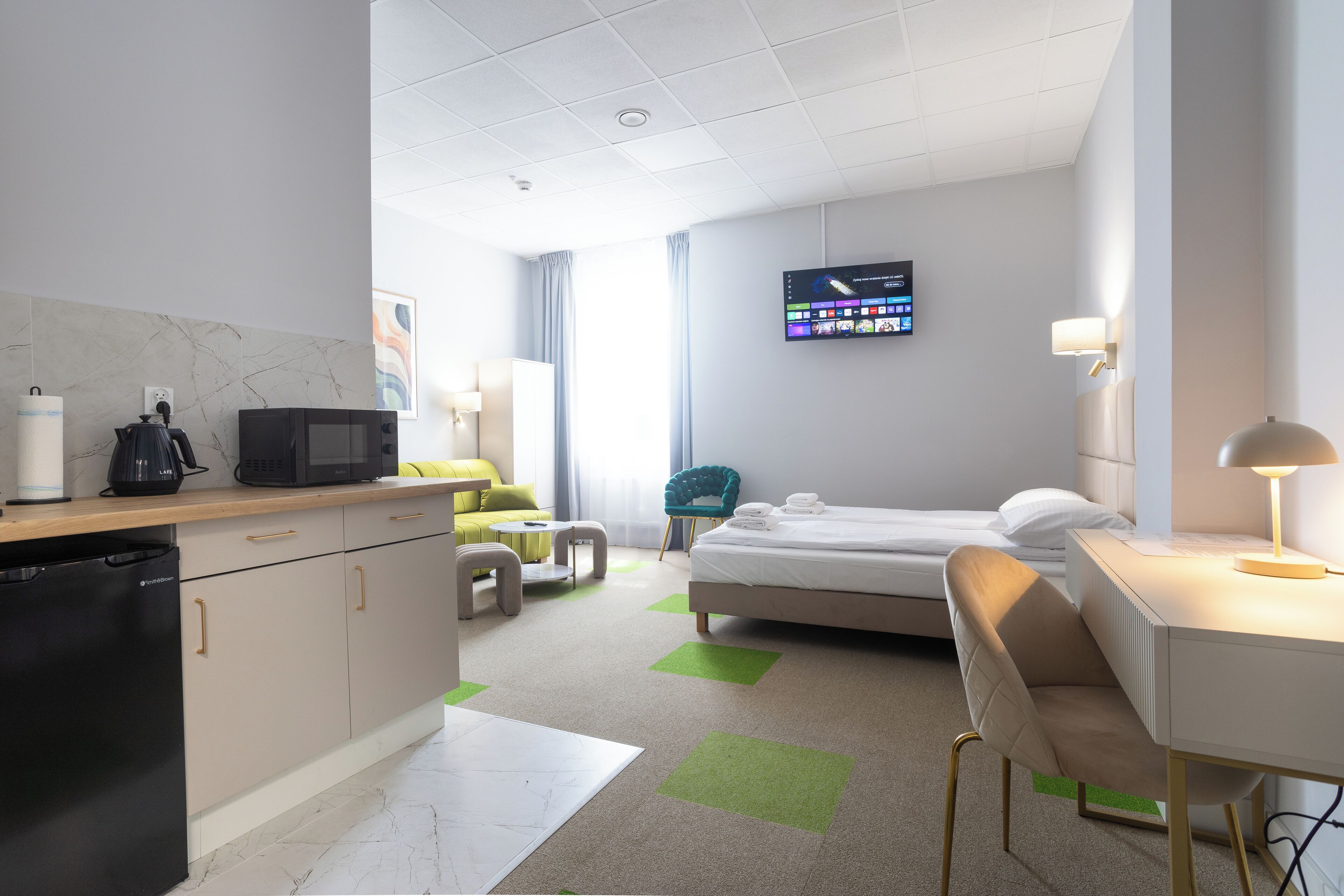 A hotel room with a bed, a desk, a chair, a TV, a microwave, and a small kitchenette.