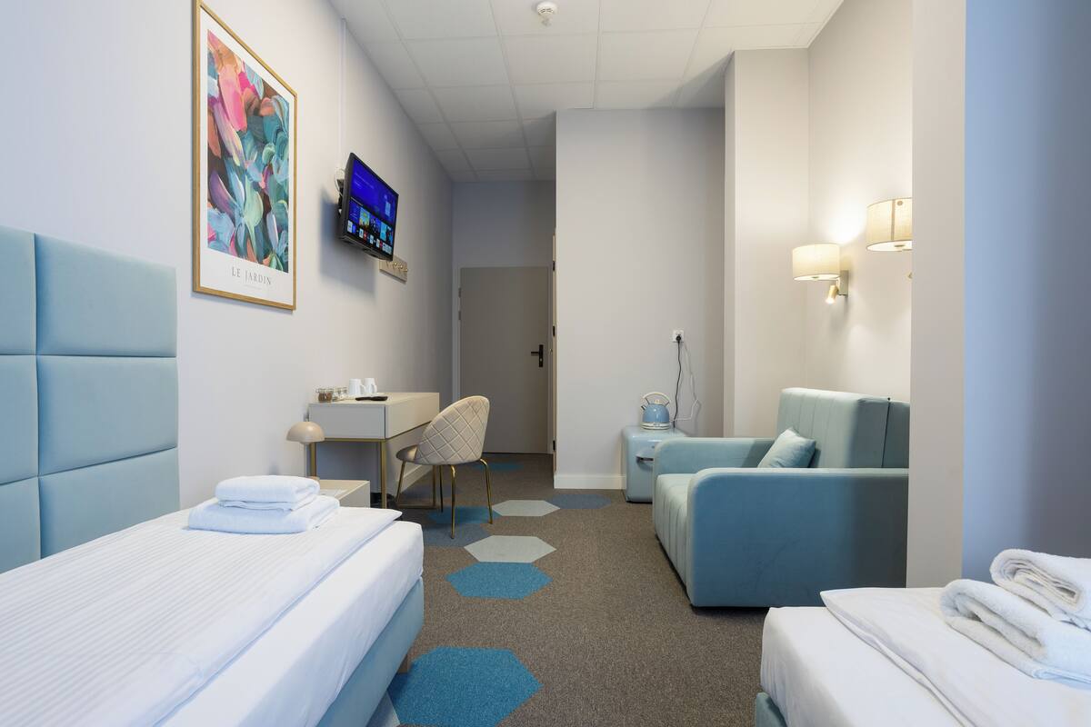 A hotel room with two beds, a desk, a chair, a sofa, and a TV on the wall.
