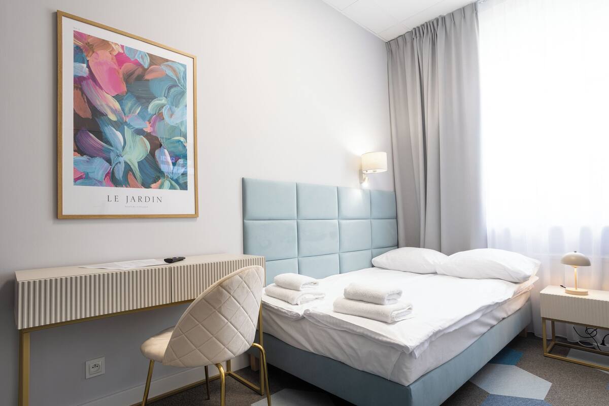 A modern hotel room with a bed, a desk, a chair, a framed artwork, and a window with curtains.
