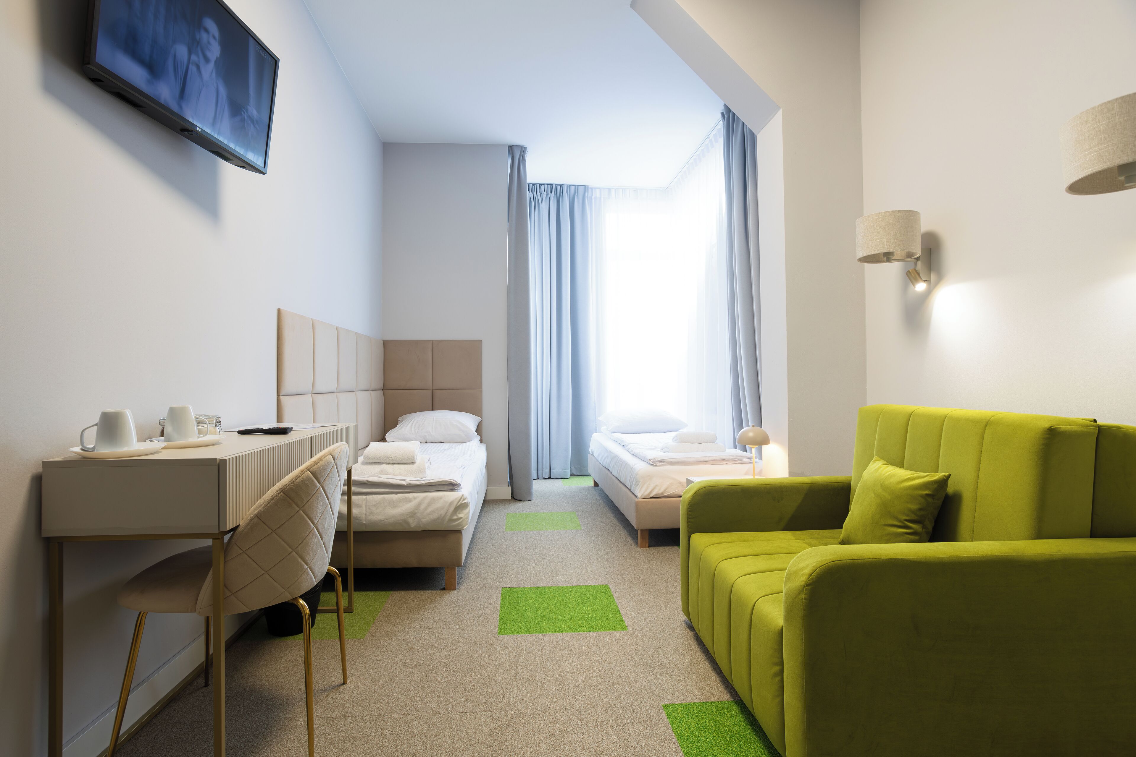 A hotel room with a desk, two beds, a green sofa, and a TV on the wall.