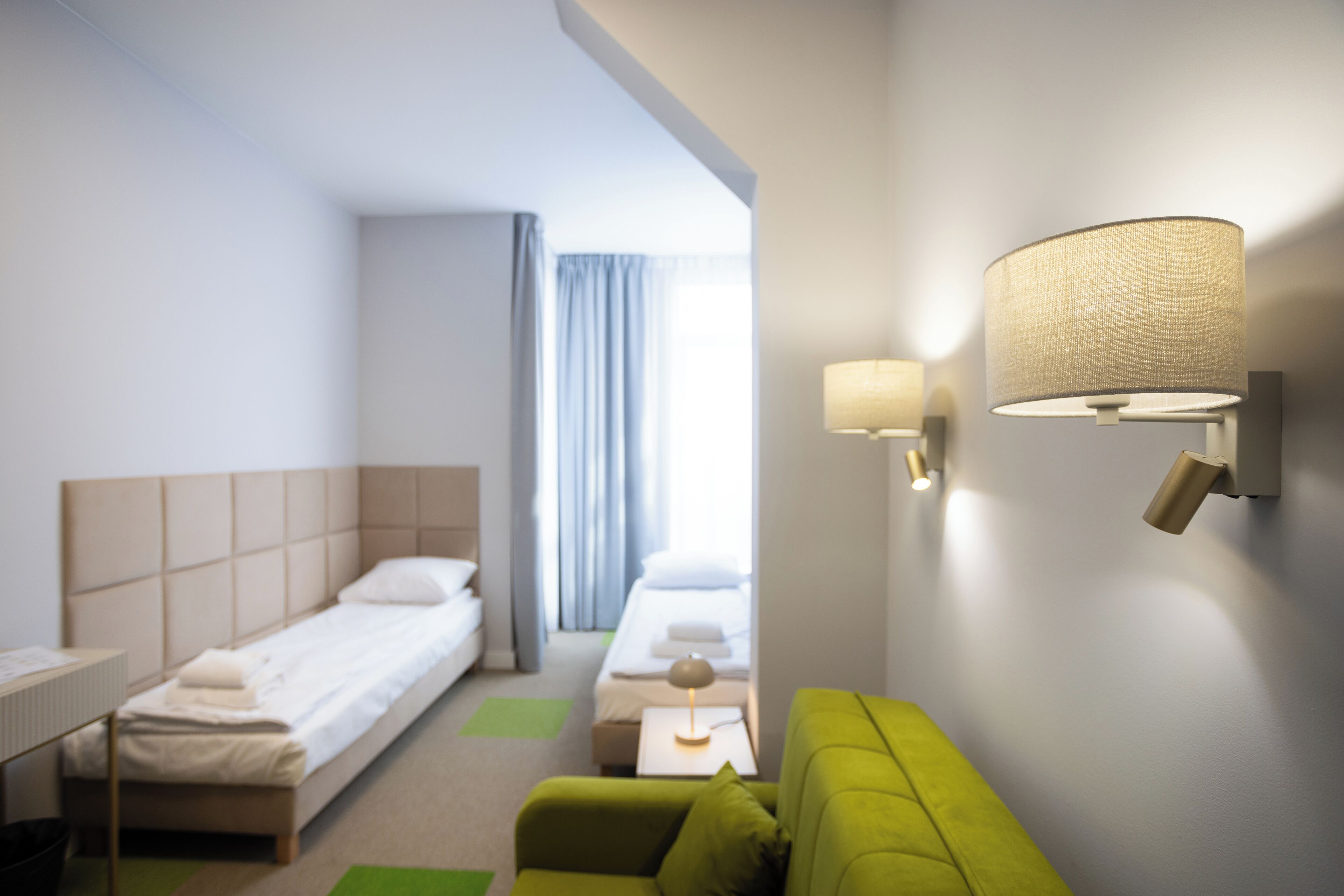 A hotel room with a bed, a green sofa, a small table, and a wall-mounted lamp.