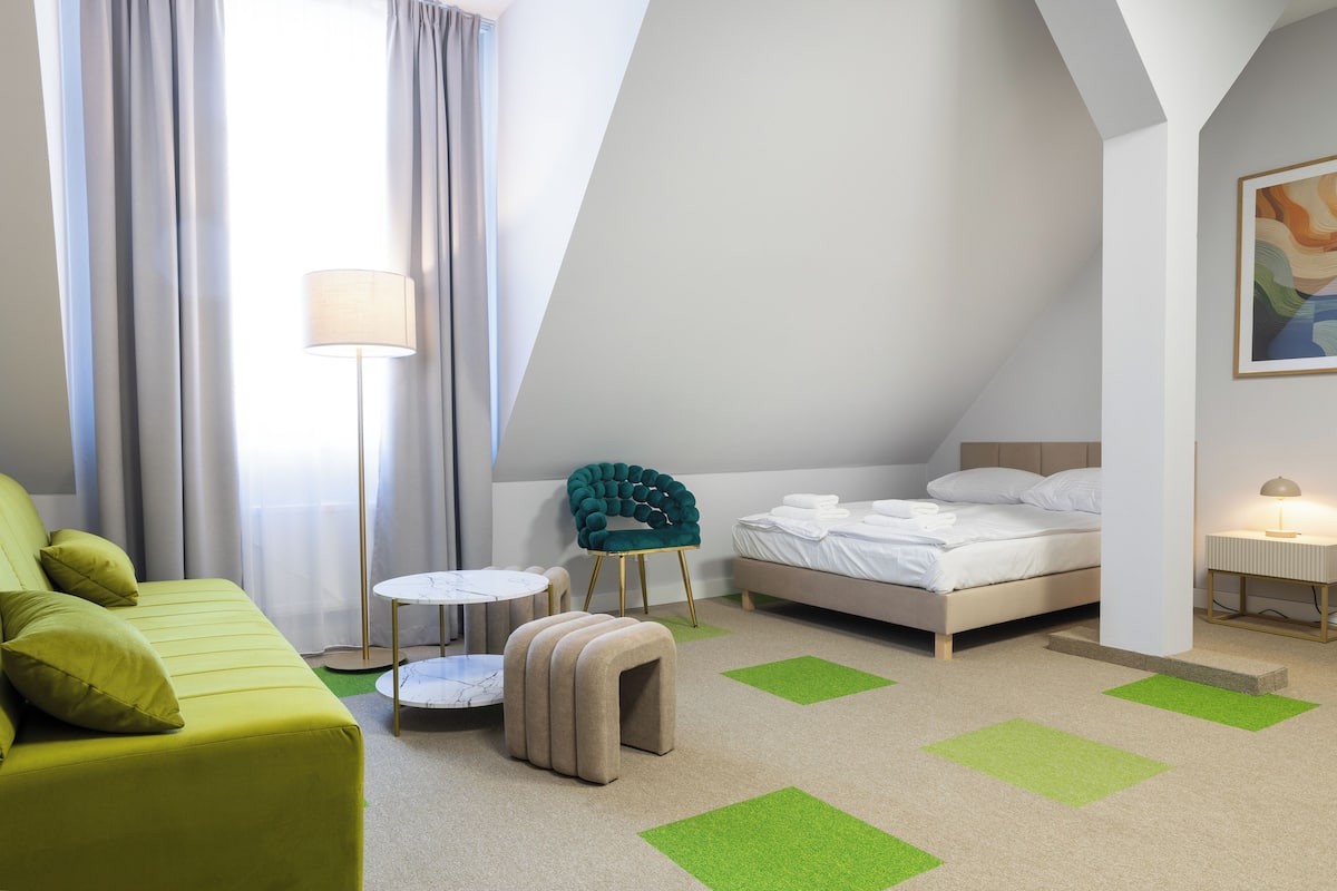 A modern hotel room with a bed, a green sofa, a small round table, a green chair, and a floor lamp.