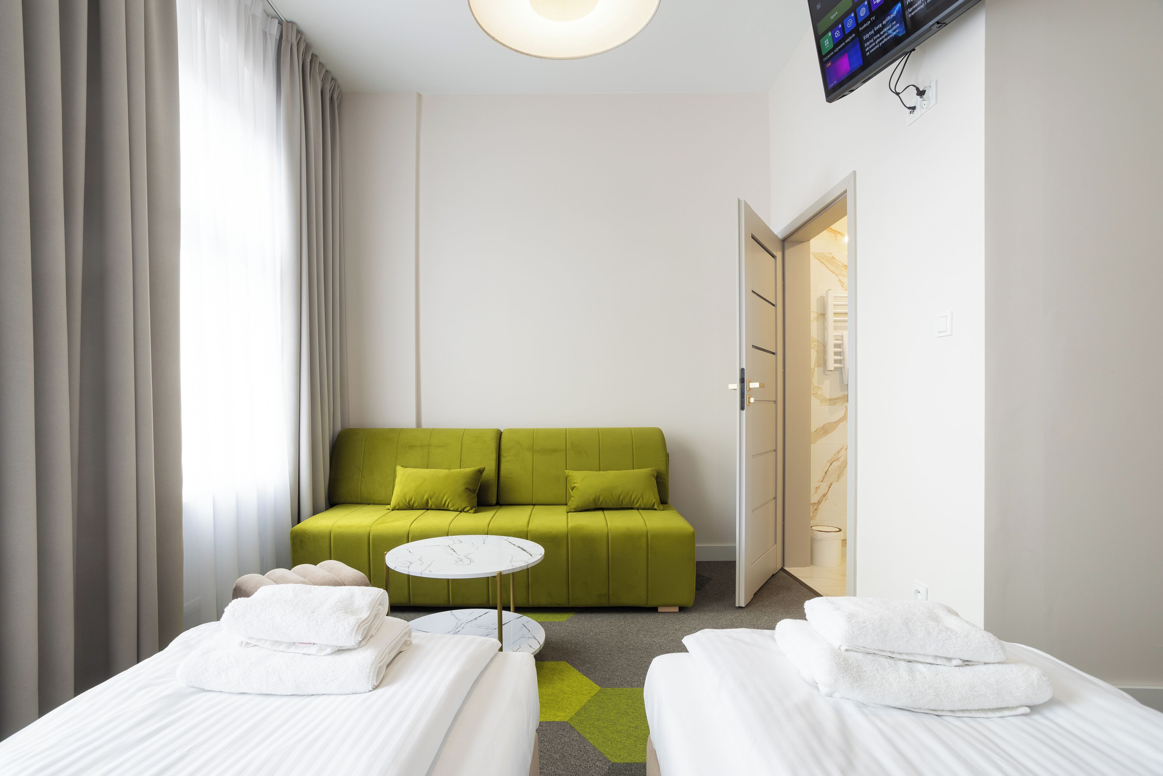 A hotel room with a green sofa, two beds with white linens, a small round table, and a TV mounted on the wall.