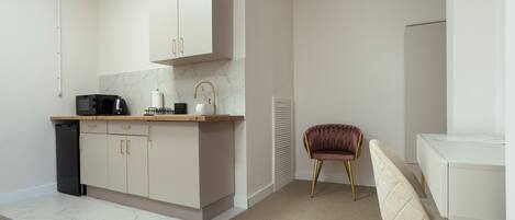 Een moderne keuken met witte kasten, een zwarte koelkast, een koffiezetapparaat en een magnetron. Er staat een bruine stoel en een witte tafel met een wit aanrecht.