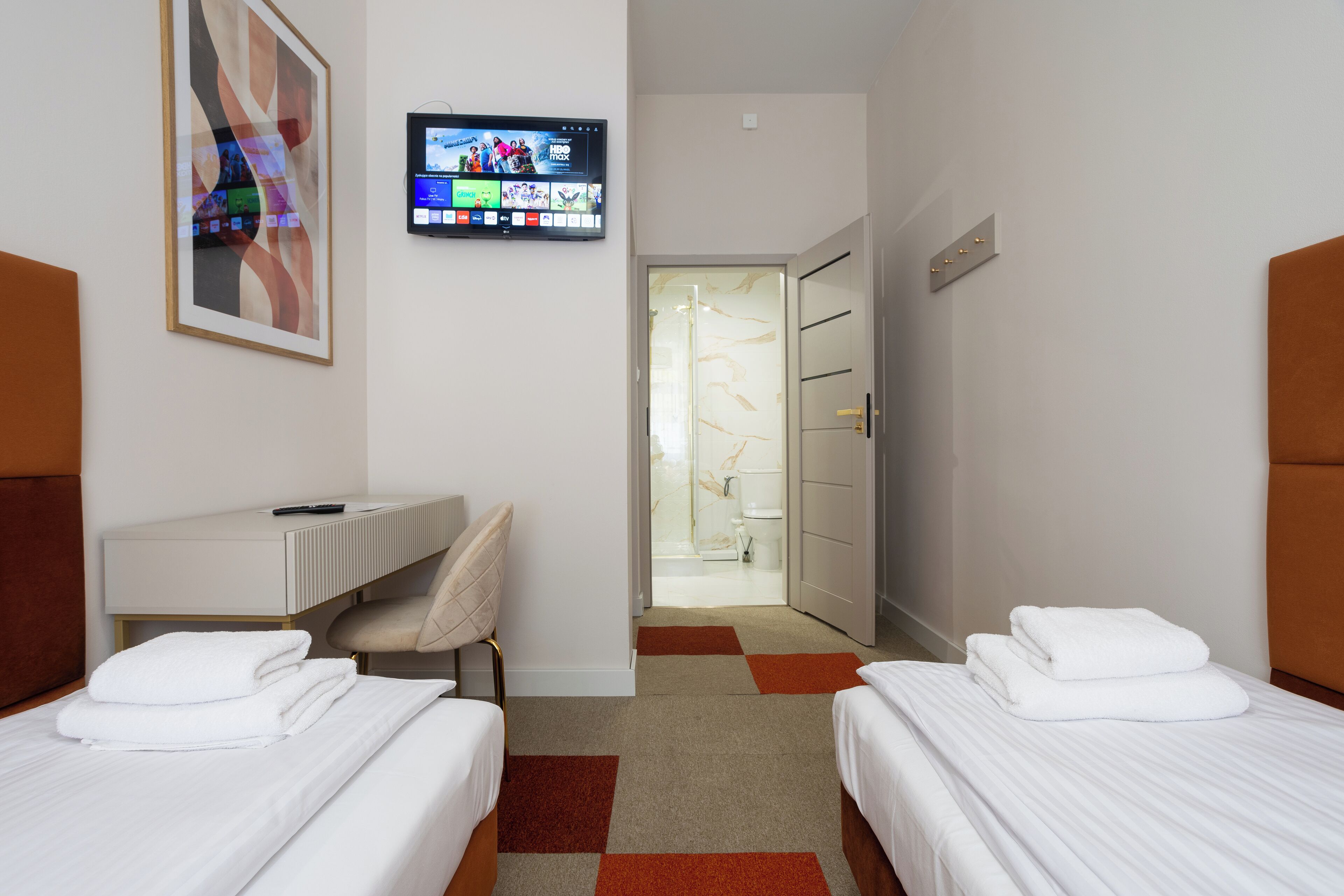 Habitación de hotel con dos camas, un escritorio, un televisor montado en la pared y un baño visible a través de una puerta abierta.