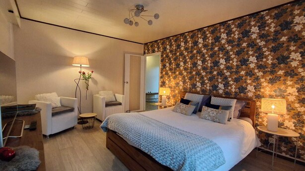 Un dormitorio con una cama grande, una zona de estar con una silla y una mesita, y una pared decorativa.