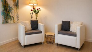 Dos sillones blancos con cojines grises, una mesita redonda pequeña, una lámpara con pantalla estampada y un jarrón con flores rojas y blancas.