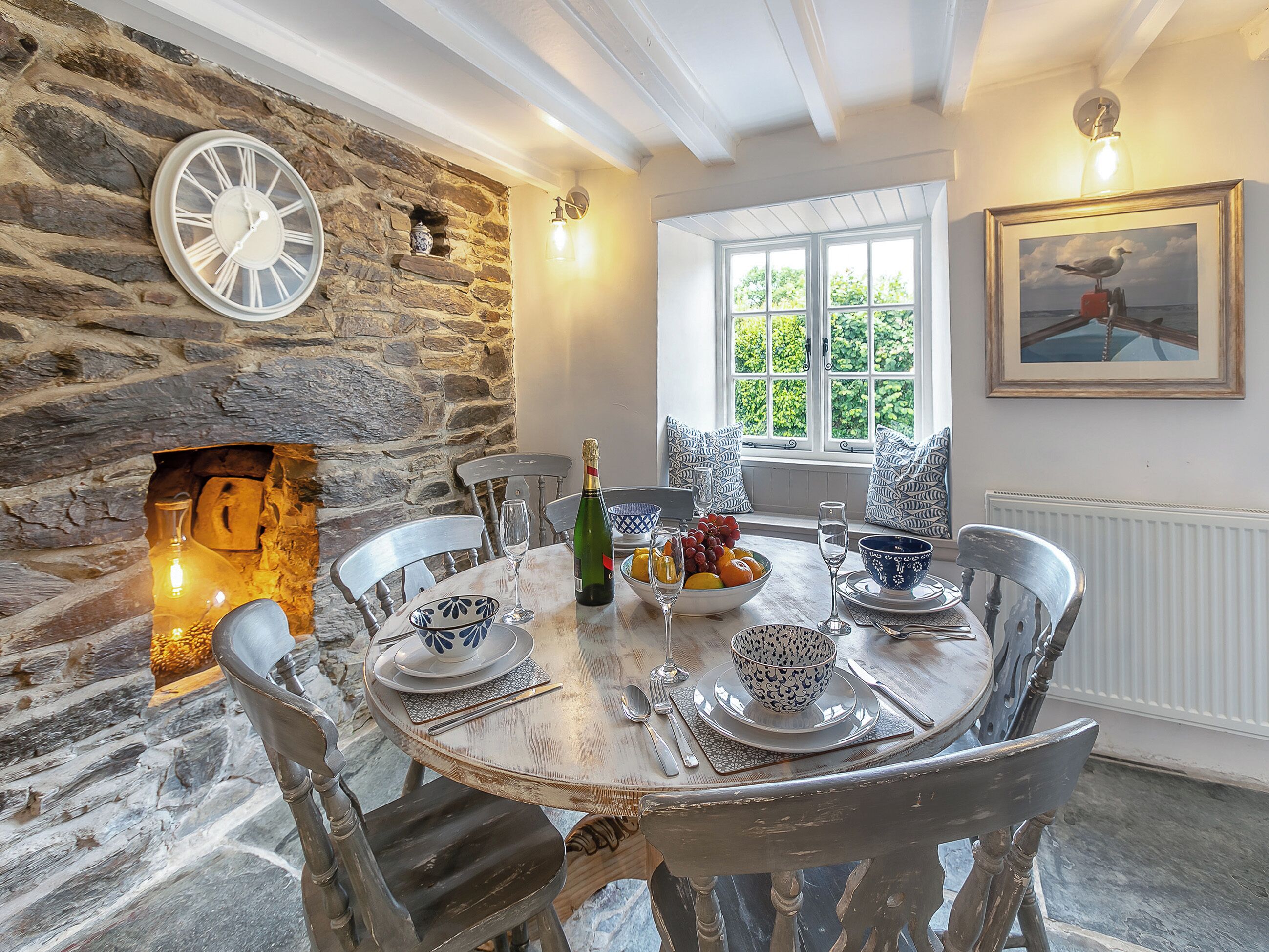 A dining room with a stone wall, a fireplace, a table set for four, and a window with a view of trees.
