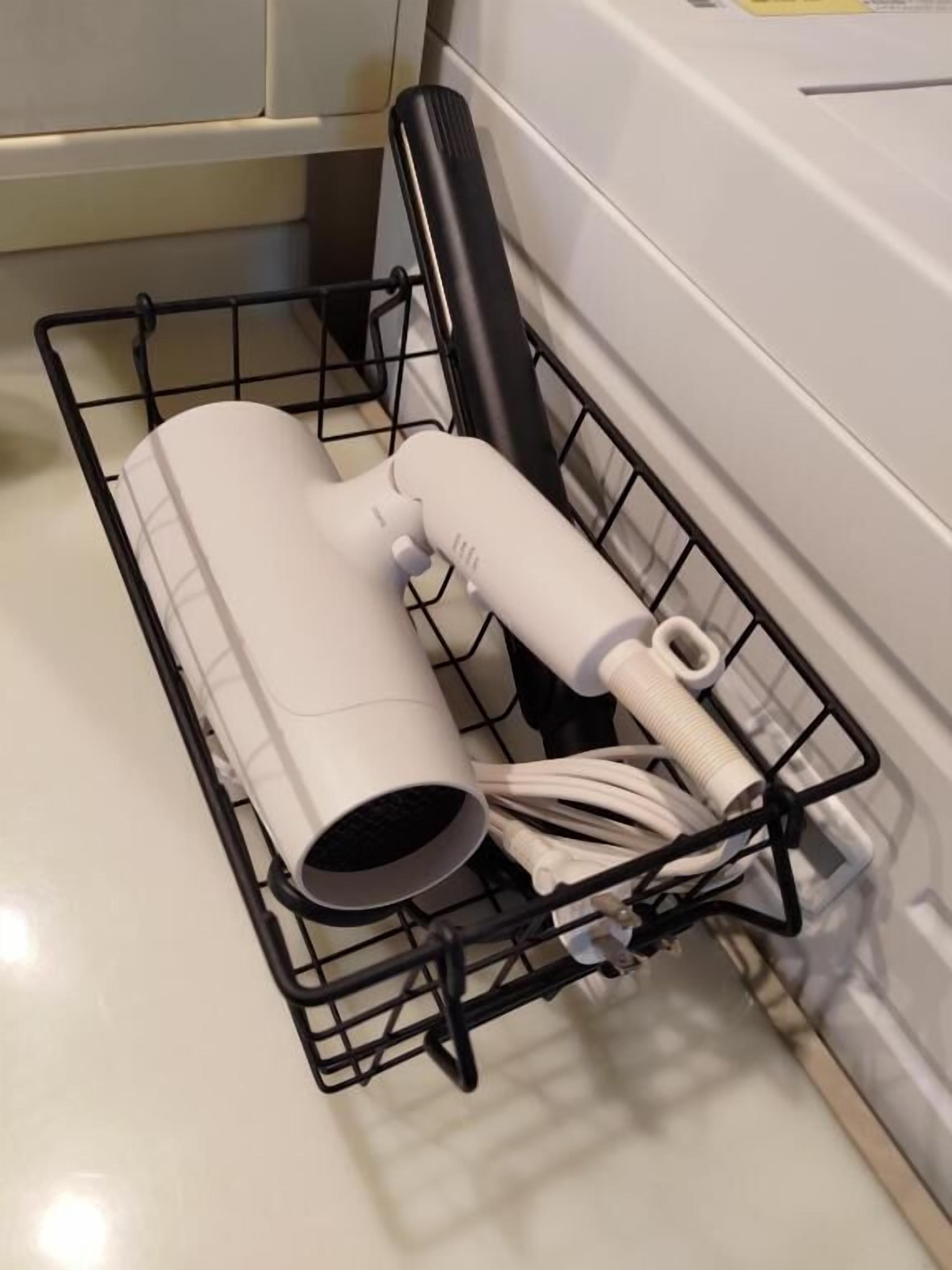 A black wire basket with a white cylindrical object and a white tube, placed on a tiled floor next to a white bathtub.