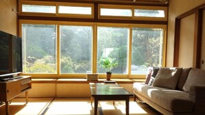 Una habitación con suelo de tatami, un sofá, un televisor y un ventanal con vistas a zonas verdes.