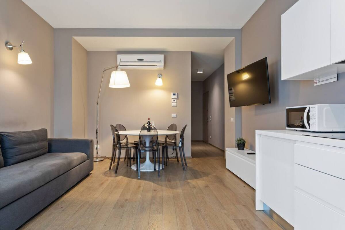 A modern living room with a gray sofa, a dining table with chairs, a floor lamp, a flat-screen TV mounted on the wall, and a microwave on a white cabinet.