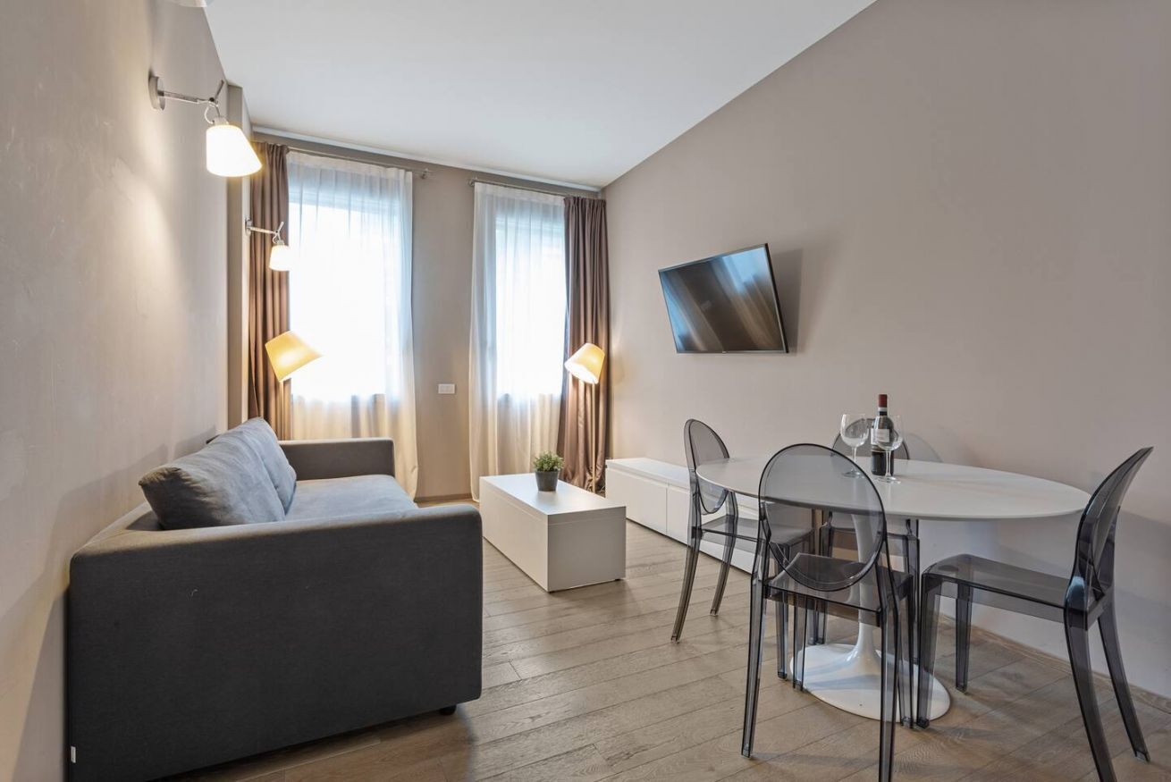 Una sala de estar moderna con un sofá gris, una mesita auxiliar blanca, una mesa de comedor de cristal con cuatro sillas, y un televisor de pantalla plana montado en la pared.