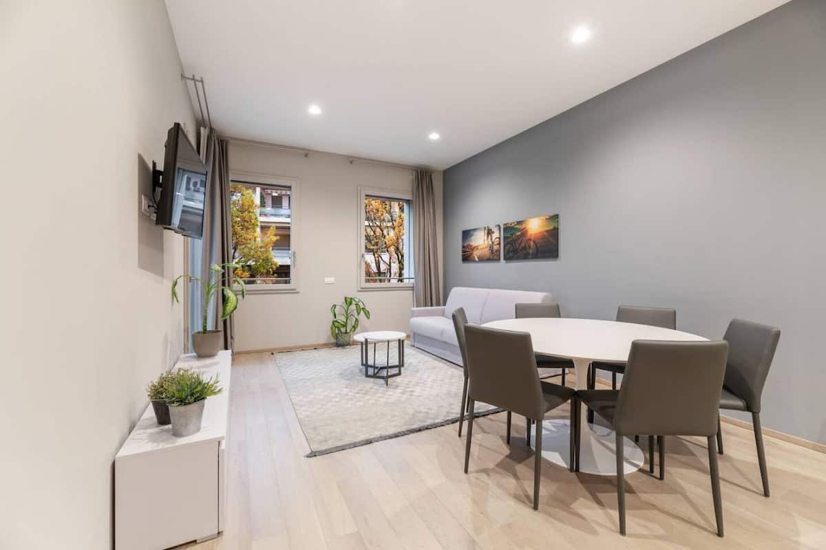 A modern dining area with a round table and six chairs, a sofa, and a TV mounted on the wall.