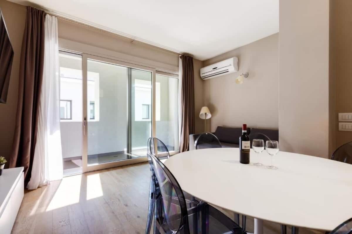 A modern hotel room with a large sliding glass door leading to a balcony, a round dining table with chairs, a bottle of wine, and two wine glasses.