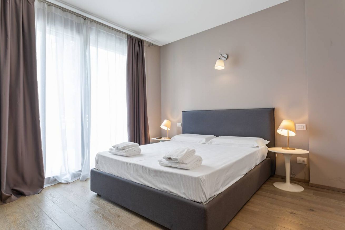 Habitación de hotel moderna con una cama grande, dos lámparas de noche y vistas a la ciudad por la ventana.