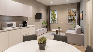 Una sala moderna con un sofá blanco, una mesita de centro y una mesa de comedor con una planta en maceta. Se observa una zona de cocina con microondas y un televisor.