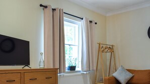 A room with a wooden dresser, a television, a window with beige curtains, a radiator, a wooden clothes rack, and an orange sofa. - 1 bedroom accommodation in Tetsworth, near Oxford (Tetsworth, near Oxford)
