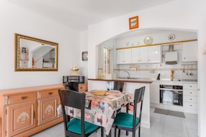 A dining area with a table set for two, a mirror, and a kitchen with white cabinets and a clock.