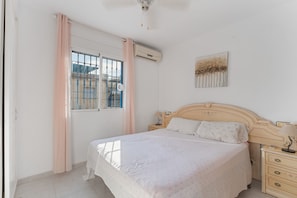 Un dormitorio con cama, mesitas de noche, ventana con cortinas, unidad de aire acondicionado y una obra de arte colgada en la pared.