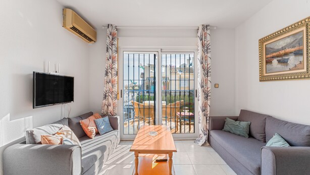 A living room with a sofa, a table, a TV, and a balcony with a view. - Apartment 'Jardin Del Mar Paraje Natural' with Lake View, Shared Pool and Air Conditioning (Torrevieja)