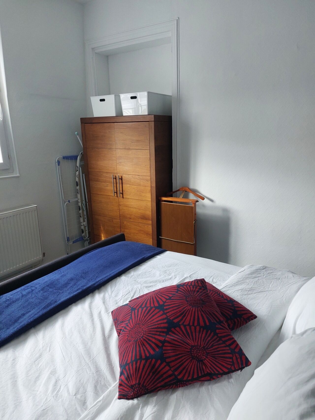 Un dormitorio con un armario de madera, una cama con ropa de cama blanca y almohadas estampadas, una toalla azul y un pequeño mueble de madera.