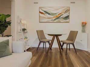 A modern living room with a white sofa, a glass-top coffee table, two armchairs, a white bookshelf, and a painting on the wall.