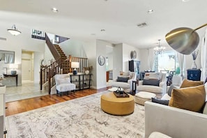 A spacious living room with a staircase, a round ottoman, and a chandelier.