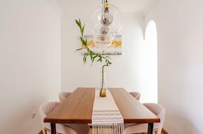 A dining area with a wooden table, two chairs, a hanging light fixture, and a wall-mounted painting.
