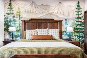 Une chambre avec un grand lit, une tête de lit en bois, deux tables de chevet avec des lampes et une fresque représentant un paysage de montagne sur le mur.