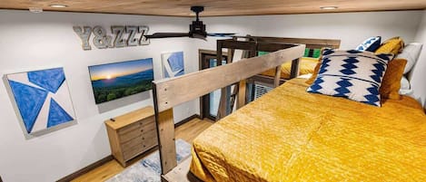 Un dormitorio con una cama, un ventilador de techo, un escritorio de madera y obras de arte en las paredes.